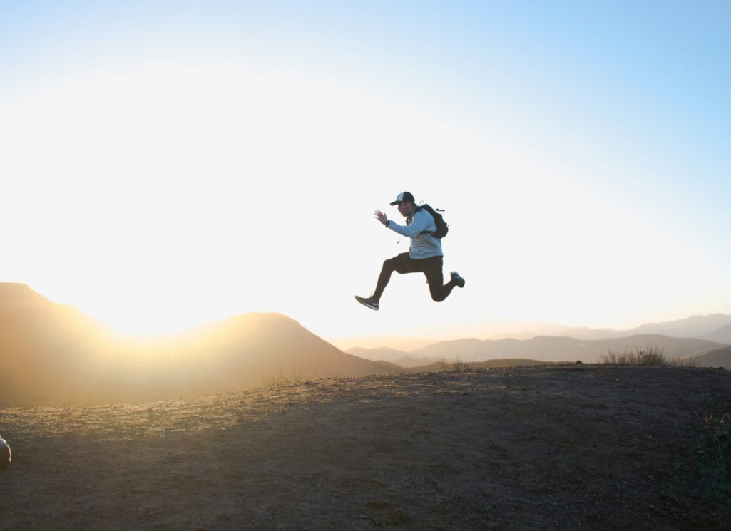 A person jumps joyfully against a sunset backdrop in a mountainous landscape.