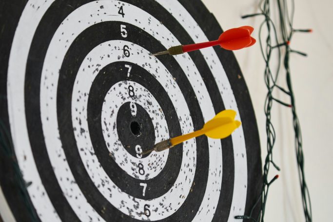 Two darts, one red and one yellow, stuck in the centre of a black and white dartboard.