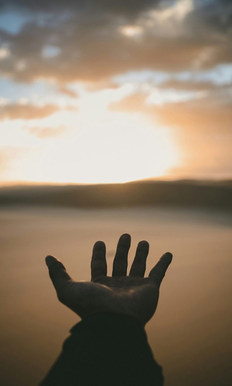 A hand reaching out towards a sunset over a tranquil horizon.